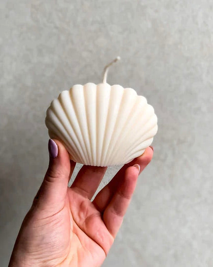 A hand with painted nails holds the SHELL CANDLE IVORY by jan-n-june—a white, seashell-shaped candle with a centered wick against a light, textured background—ideal for eco-conscious decor lovers.