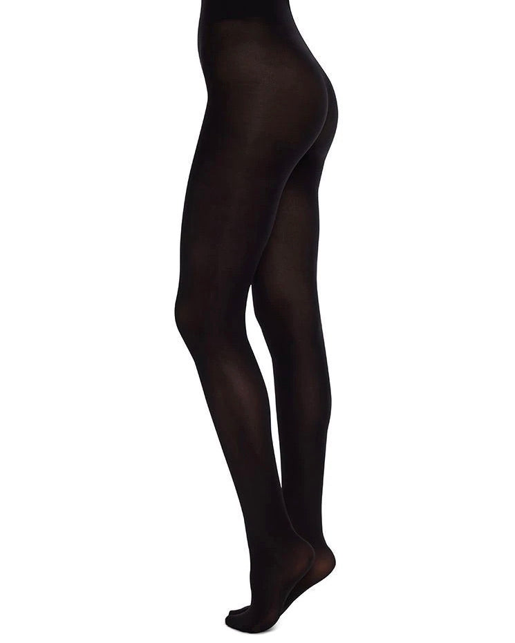 A person is wearing jan-n-junes SWEDISH STOCKINGS OLIVIA PREMIUM TIGHTS BLACK 60 DEN, made from recycled yarn. The image shows their legs and lower torso standing against a plain white background, with one leg slightly bent.