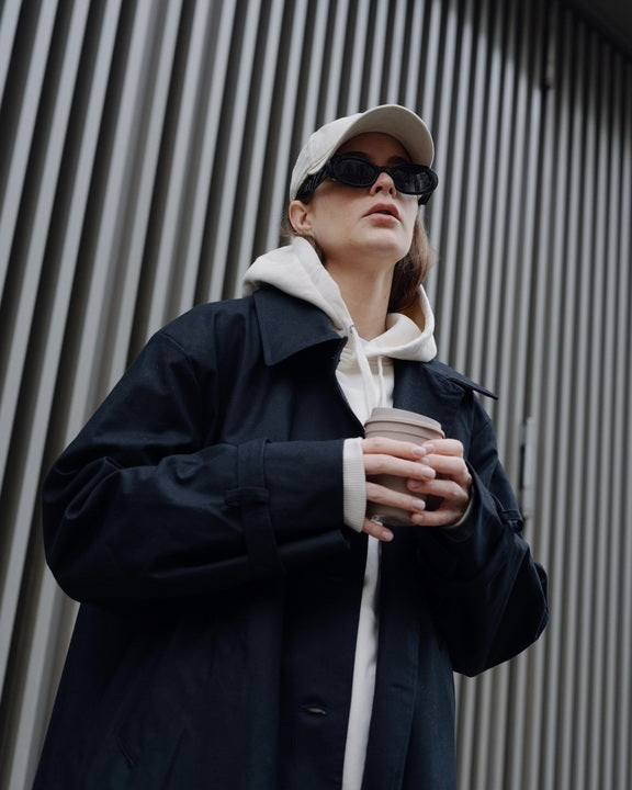A person wearing the jan-n-june COAT TAGUS BLACK with an oversized cut, paired with a white hoodie, baseball cap, and sunglasses, holds a takeaway coffee in front of a wall with vertical lines.