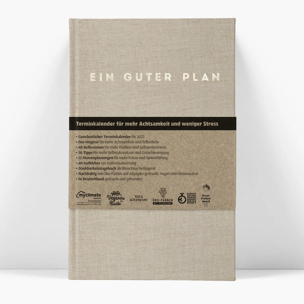 The KALENDER EIN GUTER PLAN NATUR UNDATED by jan-n-june is a beige hardcover planner with a brown label in German, highlighting mindfulness, self-love, stress reduction, eco-friendly production, and certification icons.