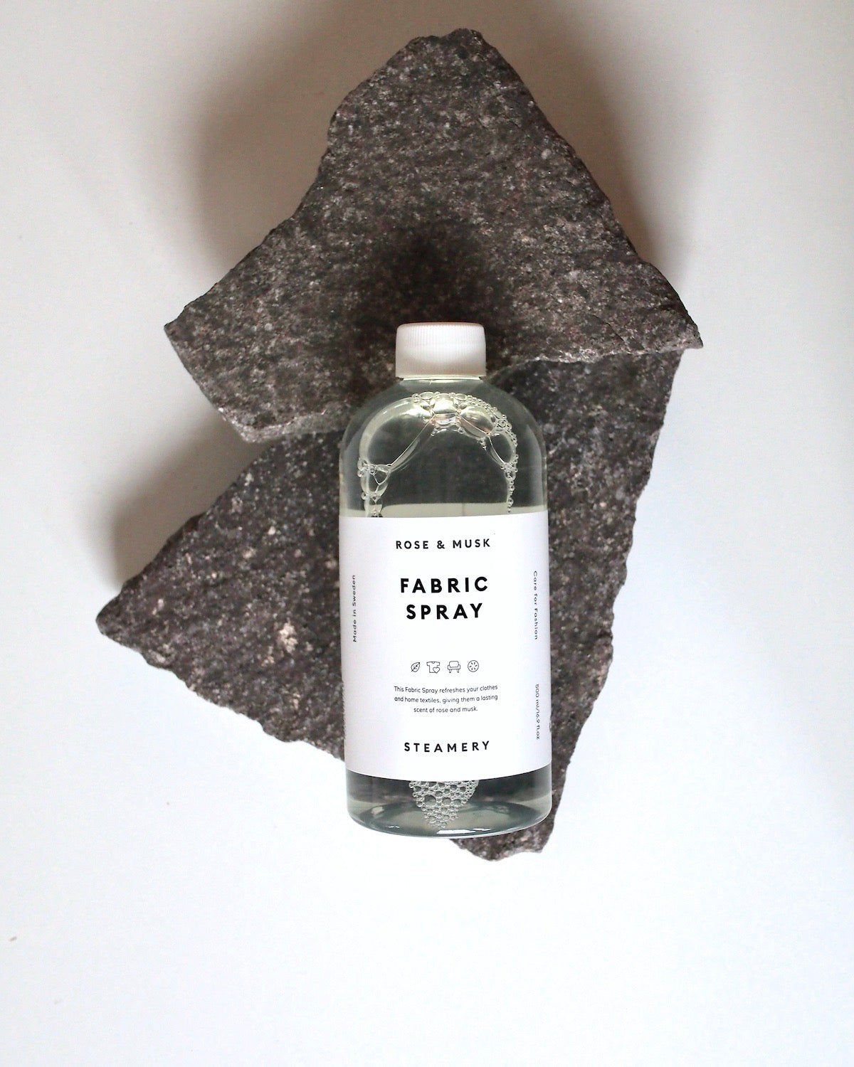 A clear bottle of jan-n-june STEAMERY FABRIC SPRAY in fresh rose & musk rests on two overlapping dark stone slabs, shot from above against a white background.