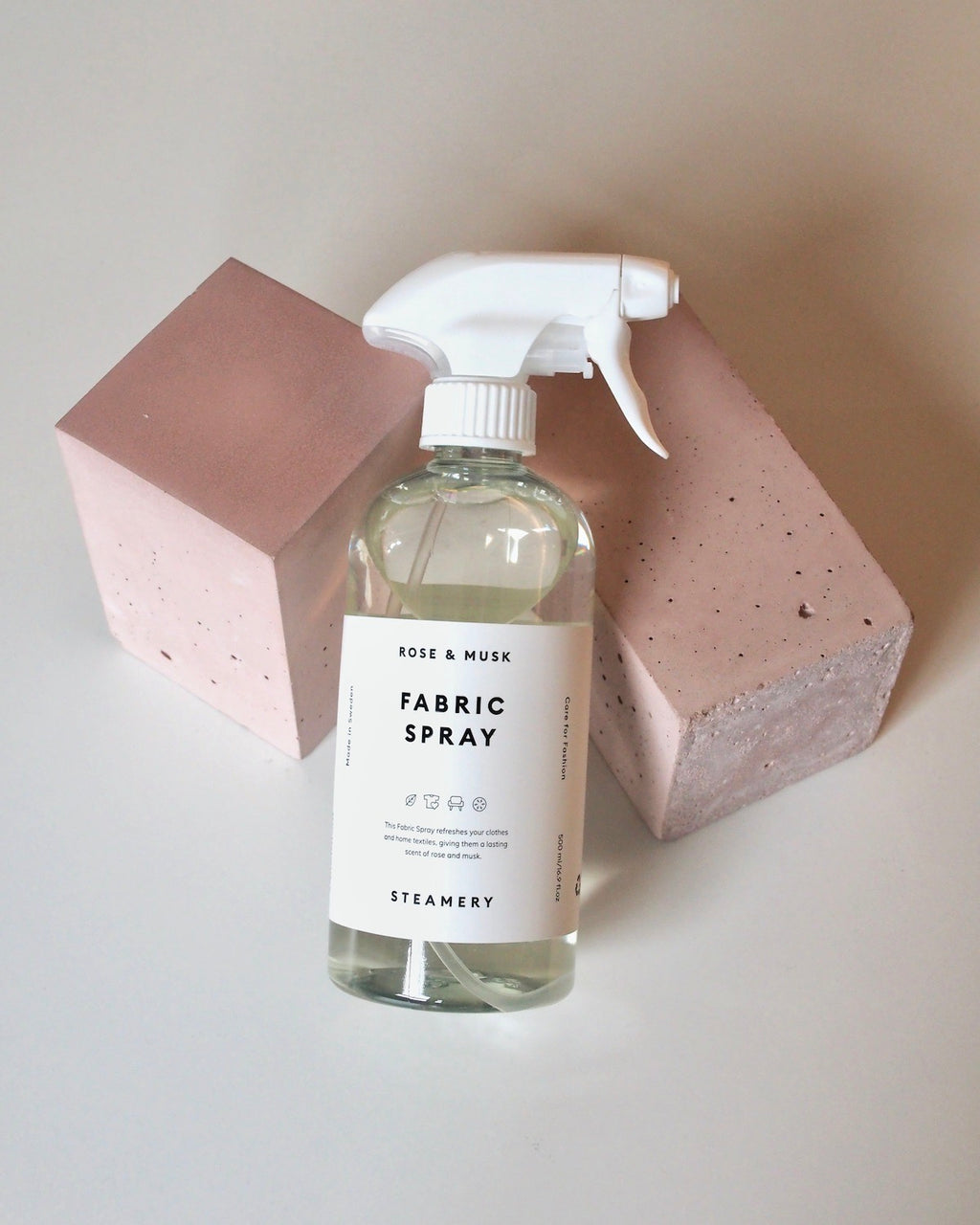 A clear STEAMERY FABRIC SPRAY bottle by jan-n-june, with fresh rose and musk scents, stands on a white surface between two light pink rectangular blocks.