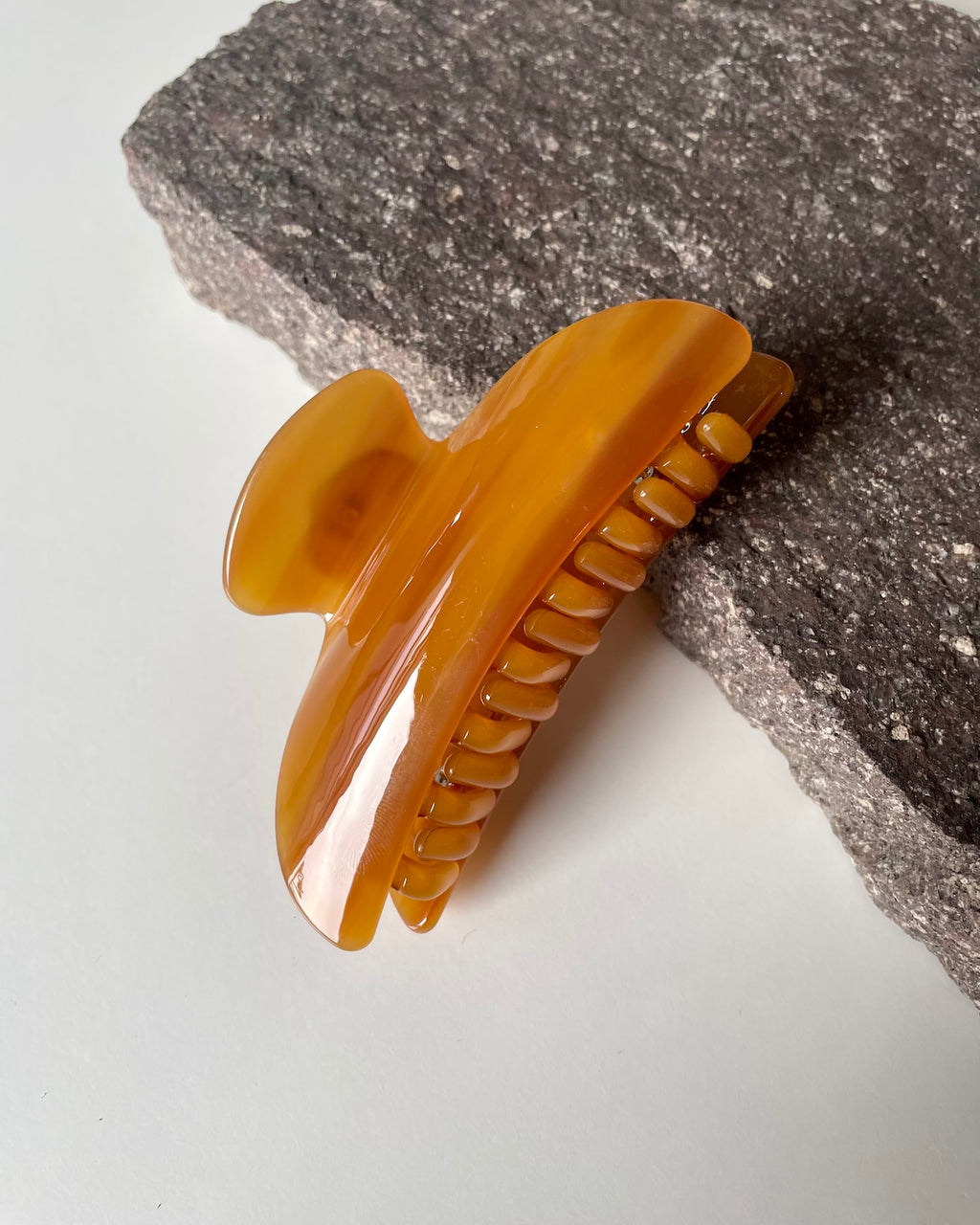 The jan-n-june MIDI HEIRLOOM CLAW COGNAC, a glossy amber acetate hair clip, sits atop a textured dark gray stone against a white background—an elegant addition to your collection.