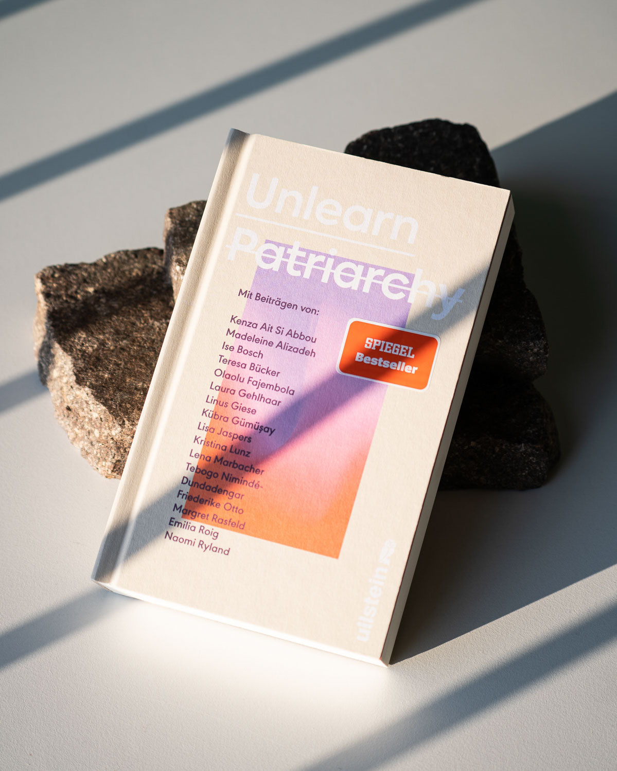 UNLEARN PATRIARCHY by jan-n-june, an anthology featuring contributors on the cover, rests on dark stone in natural sunlight. Focused on feminism, the book features a SPIEGEL Bestseller sticker on the front.