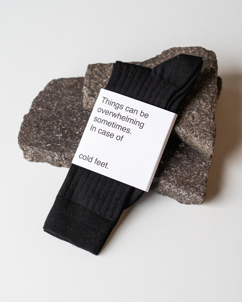 A pair of jan-n-june SOCKS BLACK rests on two dark stones, wrapped in a label that says, “Things can be overwhelming sometimes. In case of cold feet.”.