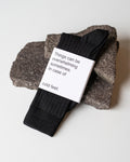 A pair of jan-n-june SOCKS BLACK rests on two dark stones, wrapped in a label that says, “Things can be overwhelming sometimes. In case of cold feet.”.