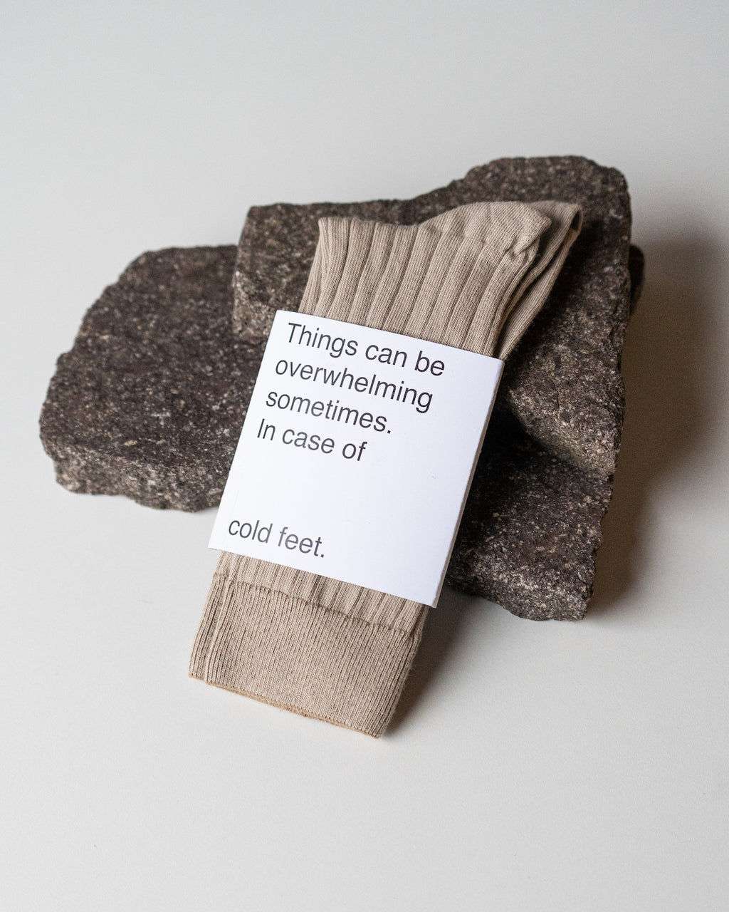 A pair of jan-n-june SOCKS ALMOND in beige organic cotton, labeled “Things can be overwhelming sometimes. In case of cold feet,” rests on two dark stones against a white background.