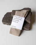 A pair of jan-n-june SOCKS ALMOND in beige organic cotton, labeled “Things can be overwhelming sometimes. In case of cold feet,” rests on two dark stones against a white background.