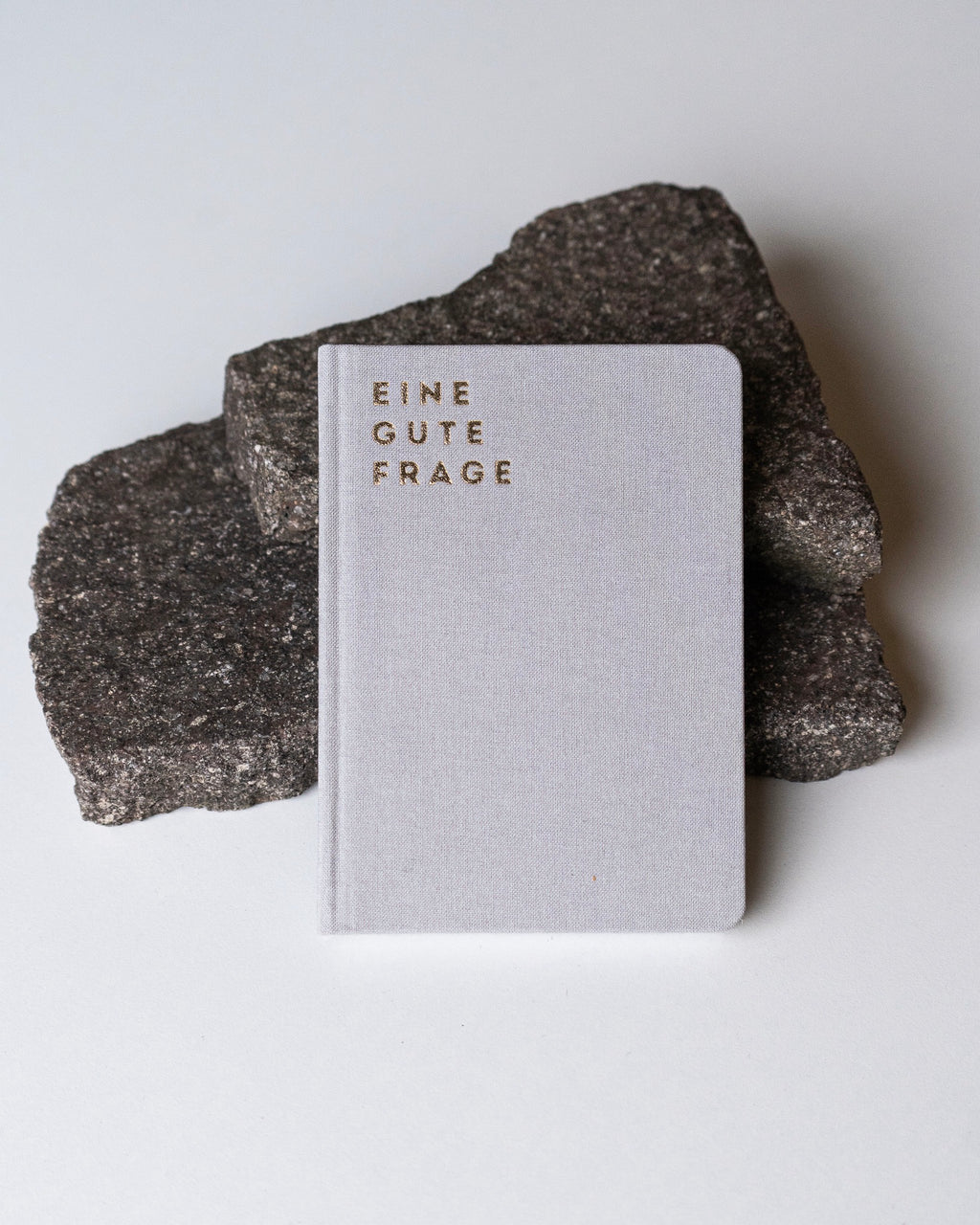 The REFLEXIONSBUCH EINE GUTE FRAGE by jan-n-june, a light grey notebook with gold embossed text, rests on dark gray stone slabs against a white background—ideal for reflecting on thoughtful questions.