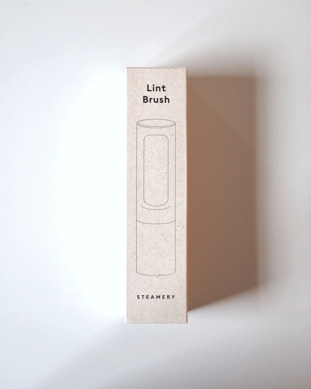A rectangular box labeled STEAMERY LINT BRUSH SAND with a simple garment brush illustration on the front and JAN N JUNE at the bottom, all on a plain white background—ideal for sustainably keeping clothes flawless.