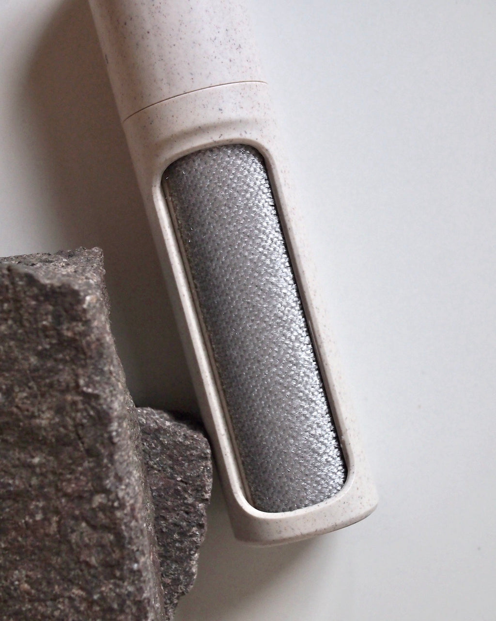 A close-up of the STEAMERY LINT BRUSH SAND by JAN N JUNE, featuring a textured metallic surface in a light beige casing, positioned beside a rough, dark grey stone on a white background.