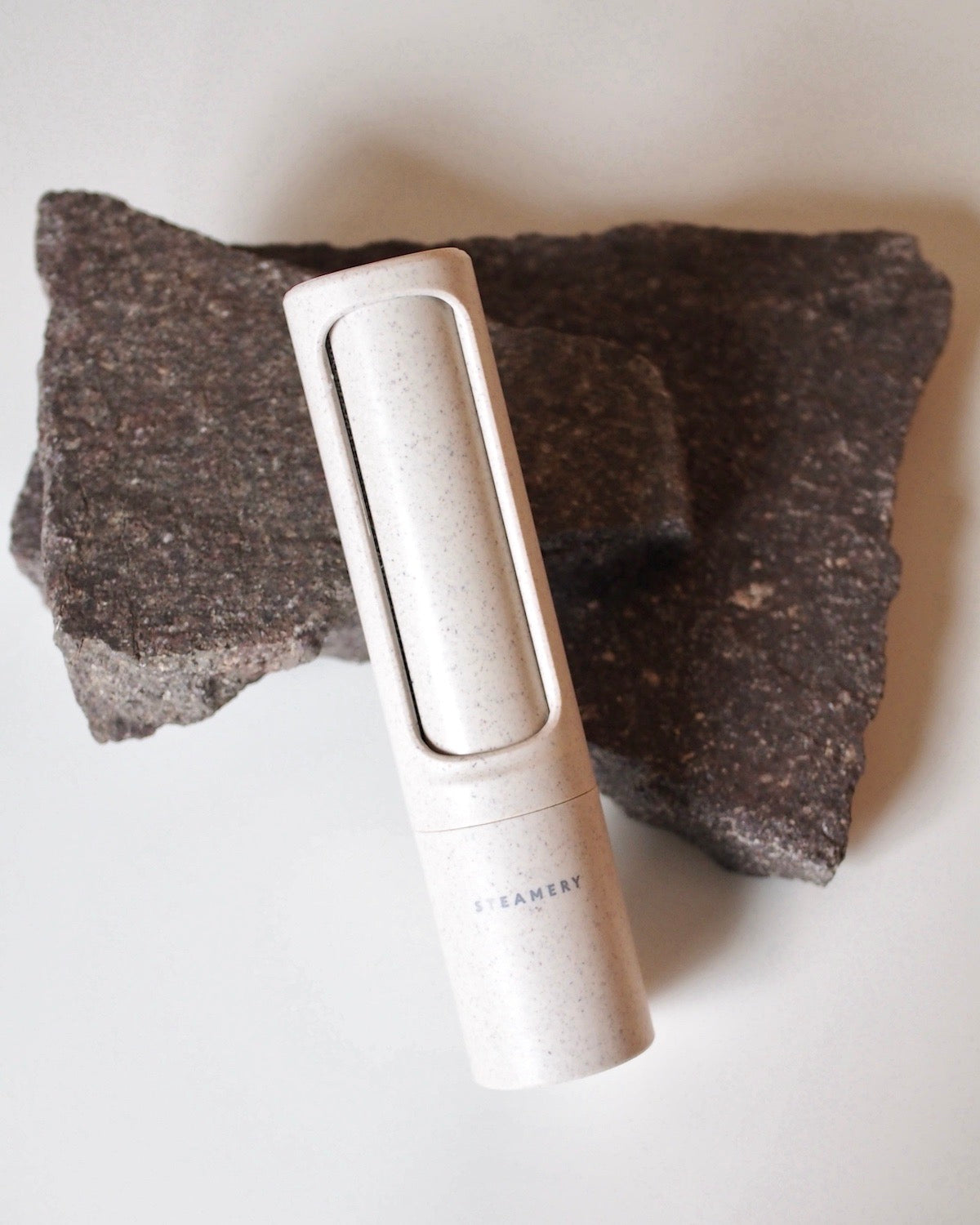A cylindrical STEAMERY LINT BRUSH SAND by JAN N JUNE, designed for flawless clothes, rests on a rough dark stone against a light background.