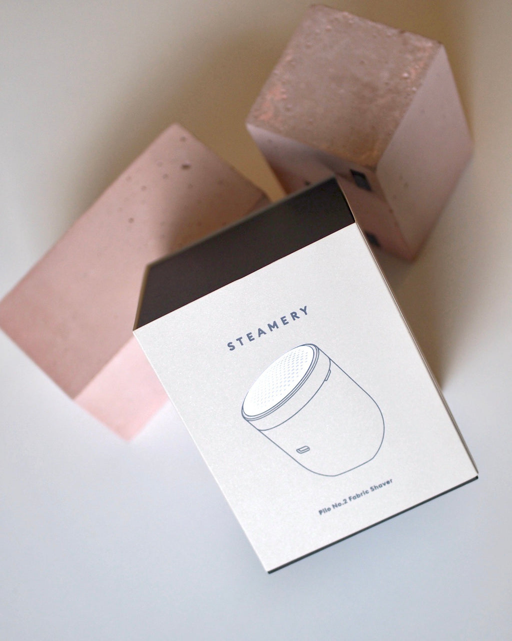 A box labeled STEAMERY PILO 2 SAND by jan-n-june, featuring a fabric shaver illustration, sits on a white surface in front of two large pink geometric blocks—showcasing its effectiveness as a lint remover.