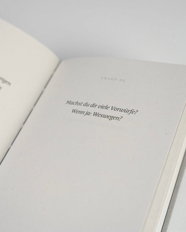 An open page of the jan-n-june REFLEXIONSBUCH EINE GUTE FRAGE displays Lena Kuhlmann’s therapeutic question in German—Machst du dir viele Vorwürfe? Wenn ja: Weswegen?—with page number 25 faintly visible.