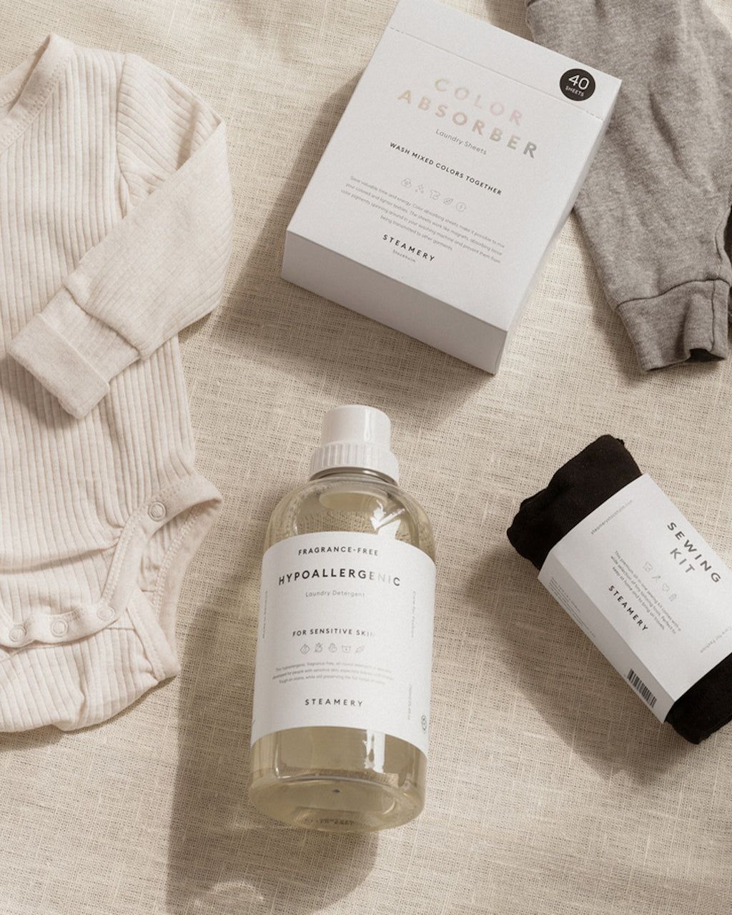 Flat lay featuring jan-n-june baby clothes, a bottle of fragrance-free hypoallergenic detergent, STEAMERY COLOR ABSORBER to prevent discoloration, and a small sewing kit, all neatly arranged on a neutral fabric background.