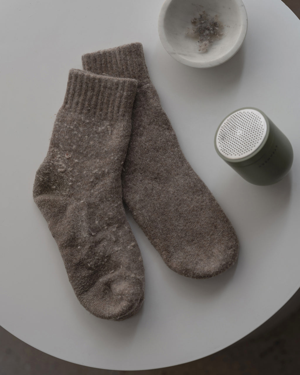 A pair of worn fuzzy brown jan-n-june socks lies on a round white table next to a small white bowl with crystals, a green cylindrical speaker, and a STEAMERY PILO 2 SAND fabric shaver for quick pilling removal.